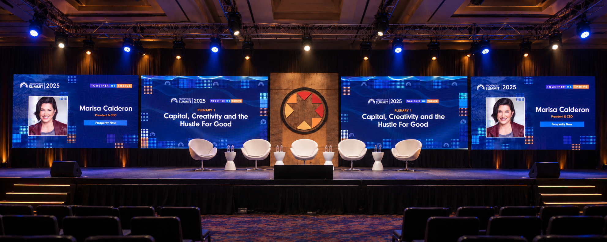 Photographic view of the Prosperity Now Summit 2025 stage with four equal landscape screens, central wood emblem and white chairs